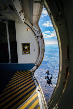 Two skydivers jump from a U.S. Army aircraft, seen from the open door with a yellow-striped floor and the Army logo, overlooking a vast landscape of mountains and clouds below.