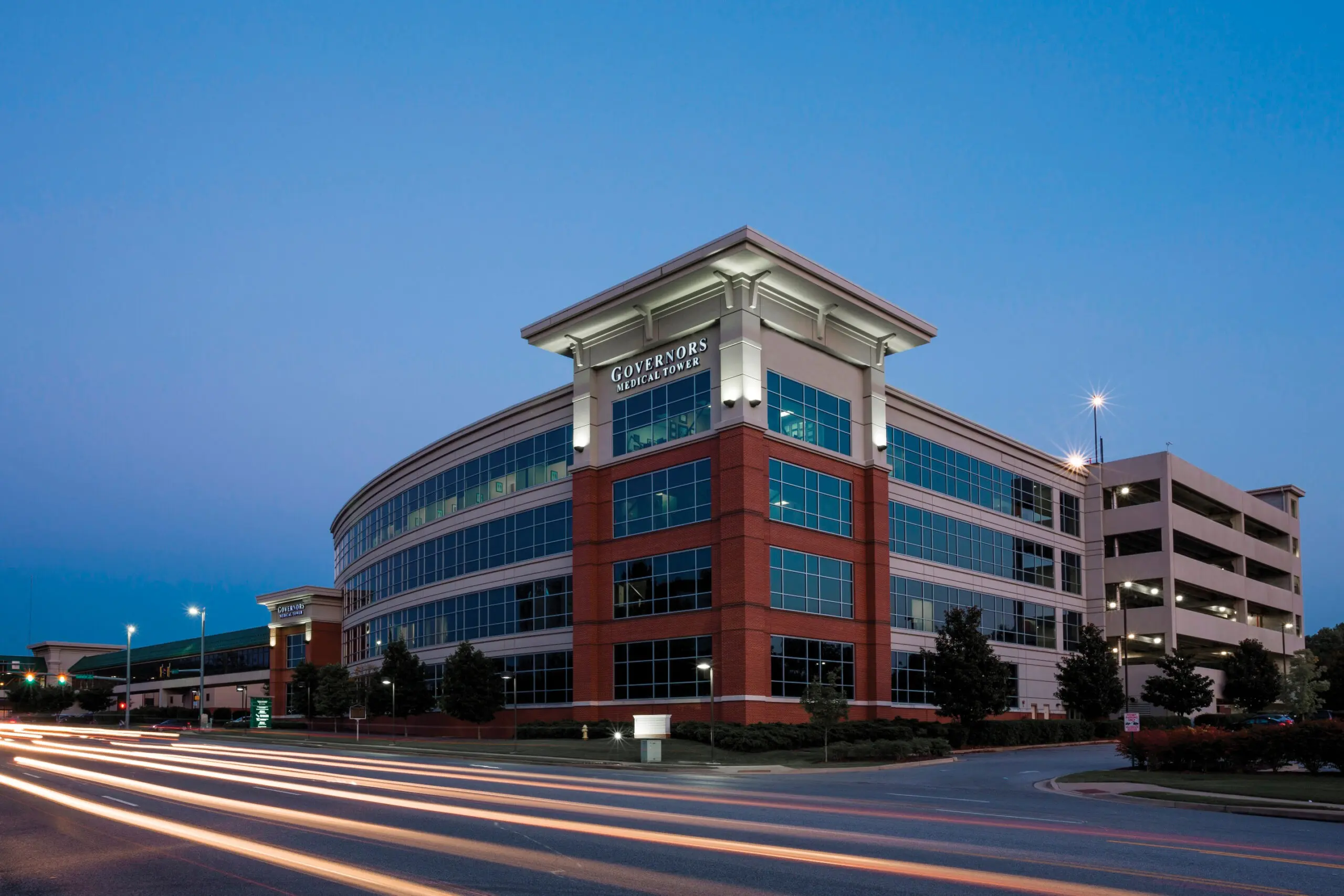 A modern office building with large windows and a sign reading Governors Centre is shown at dusk, with light trails from passing cars visible on the road in front.